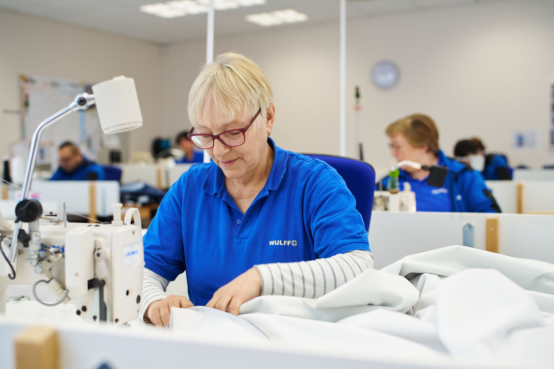 Mitarbeiterin in blauer Wulff-Arbeitskleidung näht konzentriert an einer weißen Industrienähmaschine, im Hintergrund weitere Beschäftigte in der Näherei